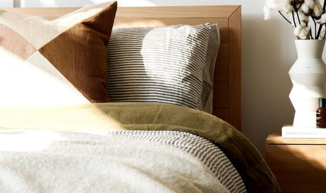 swath of sunlight shines across a well made bed and bedside table with a vase of cotton plants