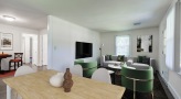 table towards bottom of photo, living room in background with tv on wall, green chairs, couch, two windows, art on walls 