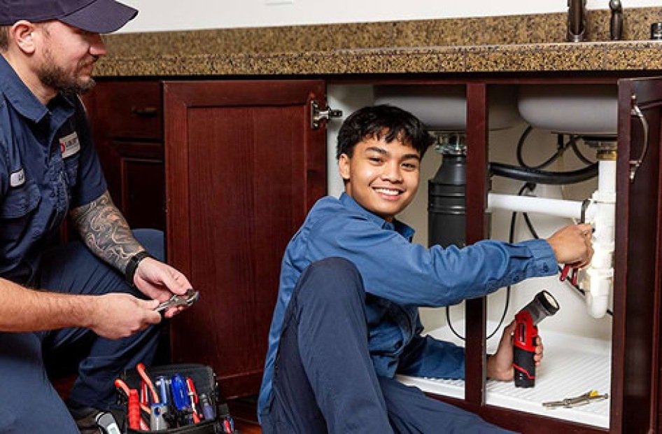 Two maintenance workers fixing plumbing under a kitchen sink.